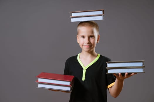 little blond boy in a black T-shirt hold stacks of books in your hands and on your head. the child is looking at the camera, isolated on a gray background.