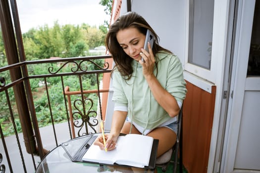 Young caucasian woman writes in a notebook with a pen in casual clothes with a phone in her hand on a summer balcony