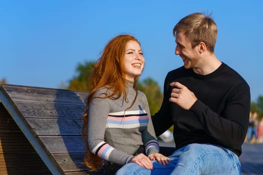 happy couple a guy and a girl with long red hair are sitting on a wooden deck in an embrace. Young man and woman of Caucasian ethnicity in casual clothes on a warm sunny day hugging