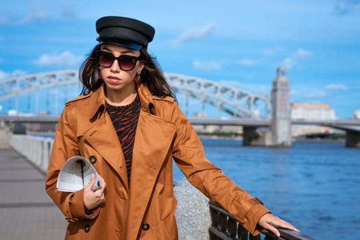 Young beautiful caucasian woman in a black cap and sunglasses in a coat on the embankment posing with a newspaper in her hand against the background of a blue sky and a bridge