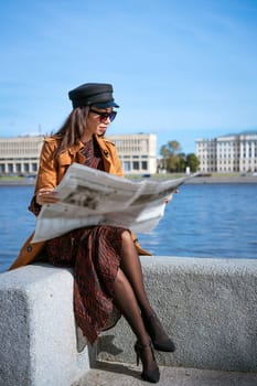 Stylish young woman of Caucasian ethnicity reads fresh newspaper in sunglasses and black cap. Beautiful brown coat standing on embankment of river on sunny day against background of sky and bridge