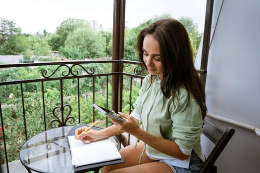 Young caucasian woman writes in a notebook with a pen in casual clothes with a phone in her hand on a summer balcony