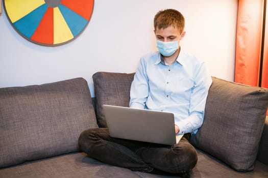 a young guy of European appearance in a light shirt, sits in a protective medical mask and works on the couch via video communication, remote work during a global pandemic