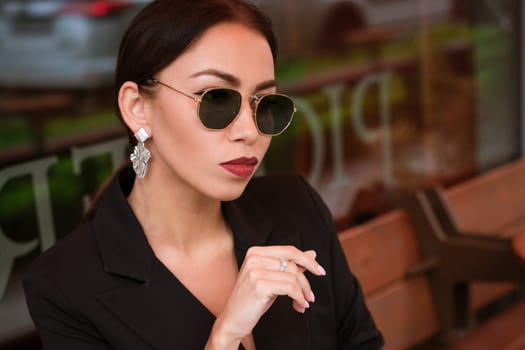 Portrait of a young business woman, brunette Caucasian appearance in a black suit and an earring sits in sunglasses in a cafe at a table. Red lipstick on lips. A business meeting