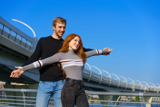 happy young couple man and woman with long red hair, stand against the background of a blue sky and a bridge in casual clothes and smile. Cheerful guy and Caucasian girl on a sunny day