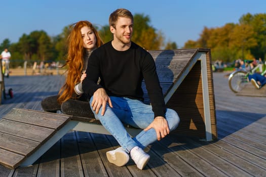 happy couple a guy and a girl with long red hair are sitting on a wooden deck in an embrace. Young man and woman of Caucasian ethnicity in casual clothes on a warm sunny day hugging