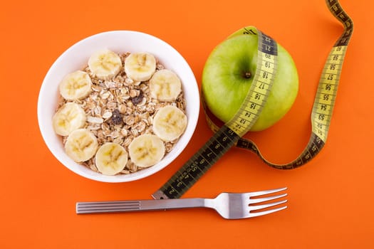 muesli with banana in a white plate on an orange background, a fork and a green apple with a yellow measuring tape. Healthy food and diet concept