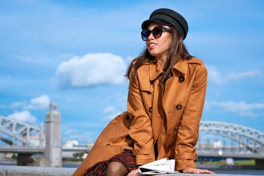 Young beautiful caucasian woman in a black cap and sunglasses in a coat on the embankment posing with a newspaper in her hand against the background of a blue sky and a bridge