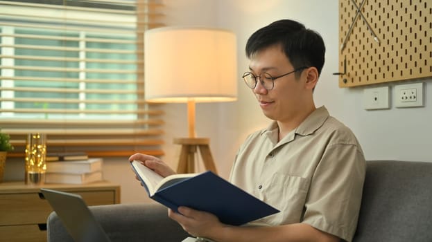 Peaceful millennial man wearing glasses sitting on sofa and reading book. Leisure and people concept.