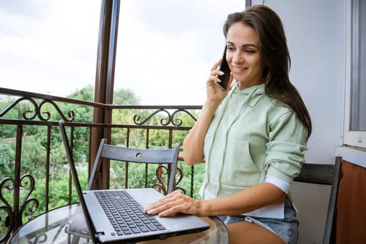 A young woman in home clothes sits on the balcony, at a table works with a phone at a laptop in the summer afternoon