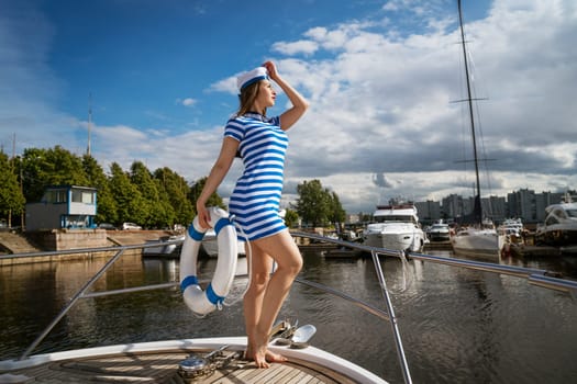 Young happy woman of Caucasian appearance in a blue striped dress standing on a yacht posing with a lifebuoy in her hand, against the background of a blue sky with clouds on a summer sunny day