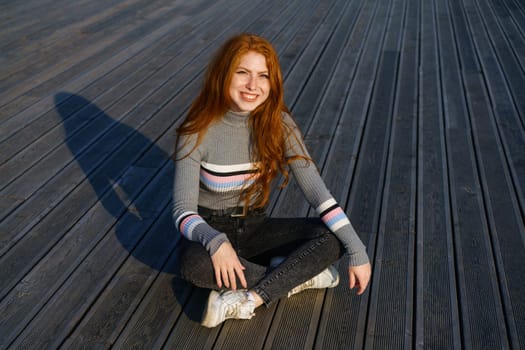 Happy young woman with long red hair of Caucasian nationality, is sitting in casual clothes in the park on a wooden deck on a sunny day and smiling