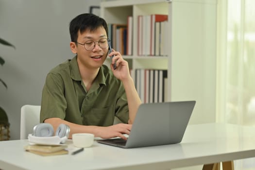 Positive millennial man in glasses talking on cellphone, sitting at working desk with laptop computer.