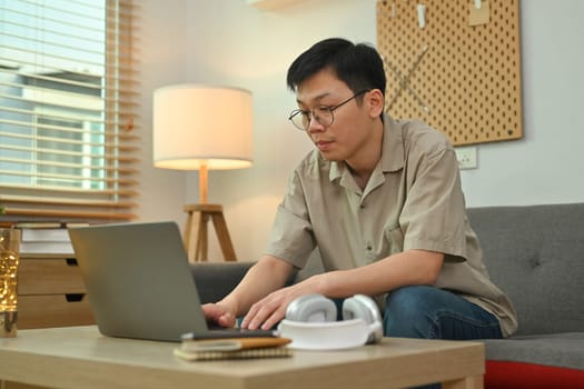 Young asian man sitting on couch and using laptop computer. Freelance, remote work and e-learning concept.