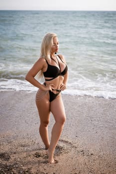a beautiful blonde girl in an open swimsuit poses on a sandy beach, near the sea. Waves roll up on the shore, the horizon is visible in the background.