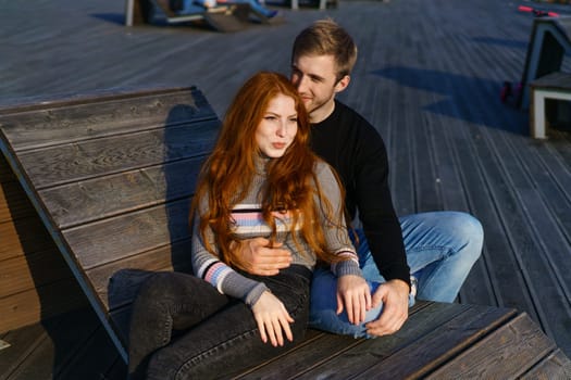 happy couple a guy and a girl with long red hair are sitting on a wooden deck in an embrace. Young man and woman of Caucasian ethnicity in casual clothes on a warm sunny day hugging