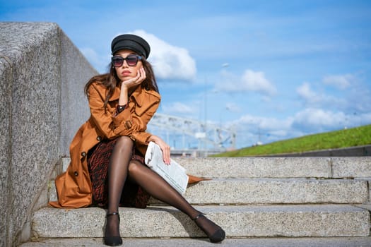 Young woman in sunglasses of Caucasian ethnicity sits on the steps on the embankment in a black cap and brown jacket with a newspaper in her hand