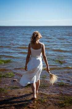 A blonde girl in a white dress walks barefoot into the sea, holding a bouquet of flowers