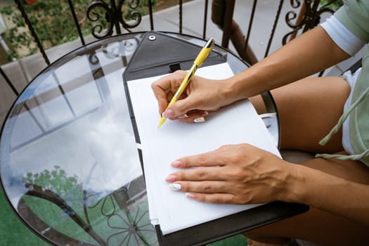 woman writes in a notebook with a pen while sitting at the table at home on the terrace