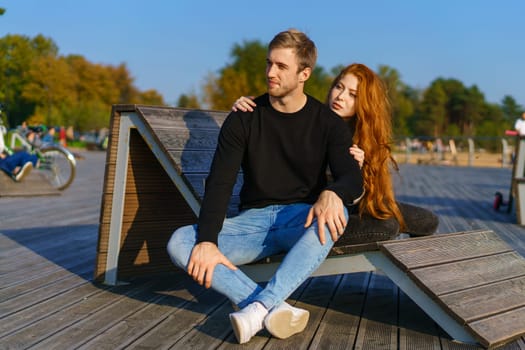 happy couple a guy and a girl with long red hair are sitting on a wooden deck in an embrace. Young man and woman of Caucasian ethnicity in casual clothes on a warm sunny day hugging