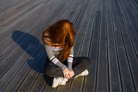 sad redhead girl sits on a wooden background in the park on a sunny day alone. Young woman alone. Sadness and depression concept