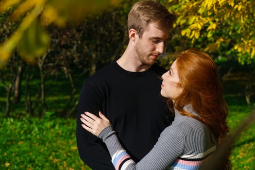 a young couple of Caucasian ethnicity, a man and a woman with red hair are standing in an embrace in a park on a sunny day against the background of trees