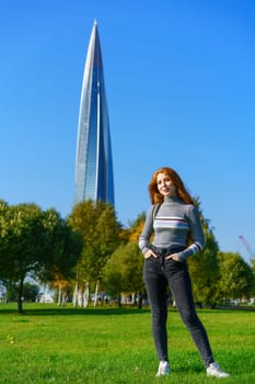 Redhead happy woman of Caucasian ethnicity with bag posing in park on green grass on sunny spring day. Cute girl in the city park against the background of green trees and blue sky. High quality photo