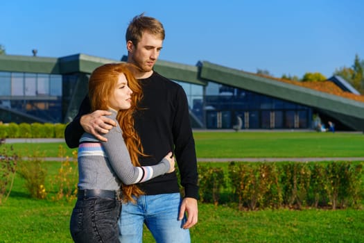 beautiful young couple, man and woman with long red hair, stand in an embrace in city park against the backdrop of modern buildings and green grass. Happy and romantic guy and girl on sunny day