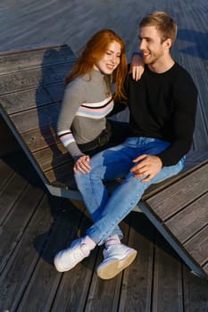 young couple of guy and girl with red hair of Caucasian appearance, in casual clothes, on a sunny day sitting in the park on a wooden bench in an embrace, happy relationship between a man and a woman