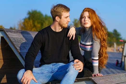 happy couple a guy and a girl with long red hair are sitting on a wooden deck in an embrace. Young man and woman of Caucasian ethnicity in casual clothes on a warm sunny day hugging
