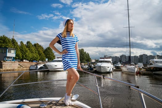Young woman of Caucasian ethnicity in a short dress with blue stripes and a cap, on a yacht, posing on a sunny day. Water entertainment concept