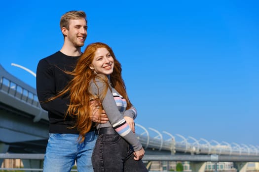 happy young couple man and woman with long red hair, stand against the background of a blue sky and a bridge in casual clothes and smile. Cheerful guy and Caucasian girl on a sunny day