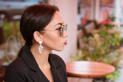 Portrait of a young business woman, brunette Caucasian appearance in a black suit and an earring sits in sunglasses in a cafe at a table. Red lipstick on lips. A business meeting
