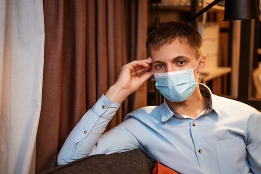 a young guy of Caucasian ethnicity in a shirt and a protective mask sits on the couch alone at home. Concept of being at home during global quarantine