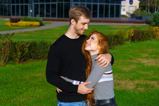 beautiful young couple, man and woman with long red hair, stand in an embrace in city park against the backdrop of modern buildings and green grass. Happy and romantic guy and girl on sunny day