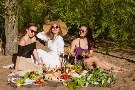 Girlfriends celebrate in the summer at a picnic. Beautiful women are having fun with alcohol in nature. Caucasian girls have fun in nature on the shore sitting on a blanket. Drink wine and eat fruit