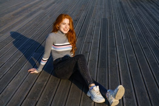 Happy young woman with long red hair of Caucasian nationality, is sitting in casual clothes in the park on a wooden deck on a sunny day and smiling