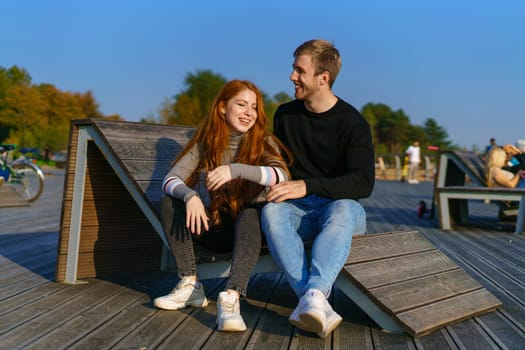 young couple of guy and girl with red hair of Caucasian appearance, in casual clothes, on a sunny day sitting in the park on a wooden bench in an embrace, happy relationship between a man and a woman