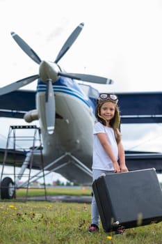 A little girl in a pilot's costume carries a retro suitcase and walks along the airfield. A child in a hat and glasses is going on a trip by plane