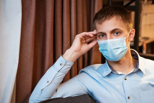 a young guy of European appearance, sitting comfortably on the couch in a medical mask. Concept to wait out the pandemic while sitting at home