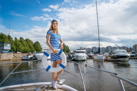Young happy woman of Caucasian appearance in a blue striped dress standing on a yacht posing with a lifebuoy in her hand, against the background of a blue sky with clouds on a summer sunny day