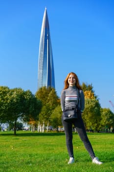 Redhead happy woman of Caucasian ethnicity with bag posing in park on green grass on sunny spring day. Cute girl in the city park against the background of green trees and blue sky