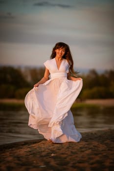 beautiful, young brunette woman in a white flowing dress on the sandy beach in the evening. full-length portrait in natural light.