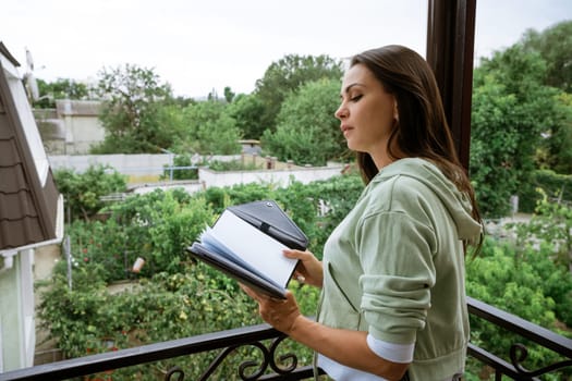 Young caucasian woman stands on the balcony with a notebook in her hand reads records