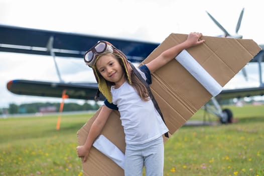 A little girl plays a pilot on the background of a small plane with a propeller. A child in a suit with cardboard wings dreams of flying.