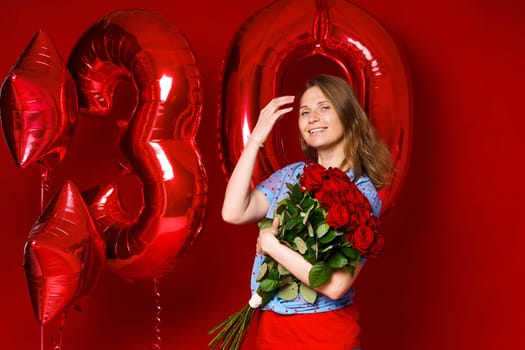 Beautiful young woman on a red background with red balloons and a bouquet of red roses. Cheerful and happy girl with bright red lipstick celebrates 30 years anniversary