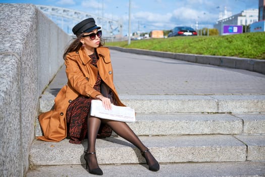 Young woman in sunglasses of Caucasian ethnicity sits on the steps on the embankment in a black cap and brown jacket with a newspaper in her hand