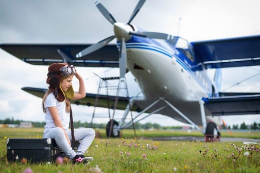 A little girl in a pilot's costume sits on a retro suitcase at the airport waiting for the departure of the flight. A child in a hat and glasses is going on a trip by plane