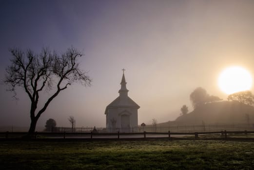 Bright sun and dark fog over small church and tree in rural countryside at dawn. High quality photo