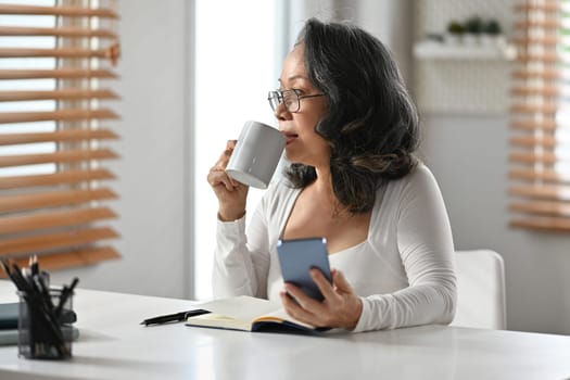 Carefree middle aged woman with mobile phone in hand drinking coffee and looking away. Retirement, technology concept.
